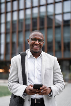 Using smartphone, messaging client, black businessman outdoor near office building, works as financial analyst, vertical. African business man smiling, prepared for client meeting, managing accounts.