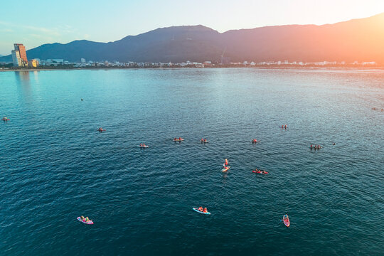 Several groups enjoy paddleboarding and small watercraft on a calm bay. A modern coastal city and majestic mountains are visible along the distant horizon at sunset