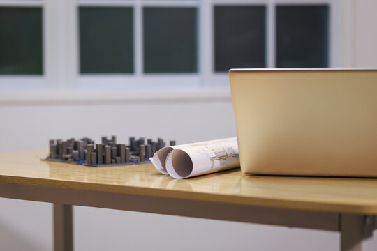 Laptop beside rolled blueprints and a small building model on a desk in a bright office workspace