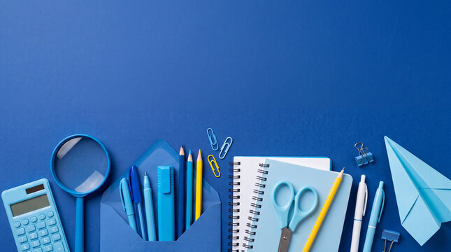 A top-down flat lay view of blue office and school stationery items including notebooks, pens, calculator, envelope, and paper plane on a solid blue background with space for text.