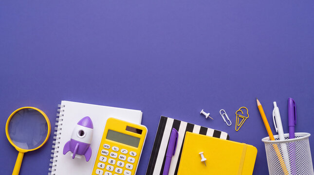 Flat lay of yellow and purple office stationery, notebooks, calculator, magnifying glass, and rocket model over a purple background with copy space.