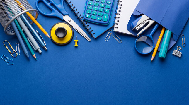 Flat lay border of blue school and office supplies, including notebooks, calculator, scissors, pens, and paper clips on a blue background with copy space.