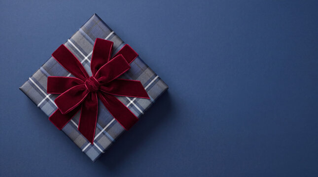 A beautifully wrapped plaid patterned gift box adorned with a rich red velvet bow, set against a dark blue background with copy space.