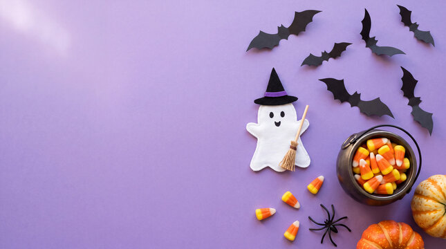 Spooky Halloween composition featuring a ghost with witch hat, flying paper bats, candy corn in a mini cauldron, spider, and pumpkins on purple background with copy space.