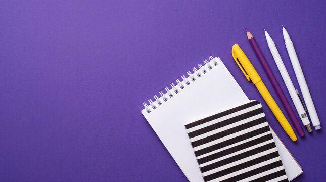 Top view flat lay of spiral notepads with colorful pens arranged neatly on a vibrant purple background, perfect for education and office concepts.