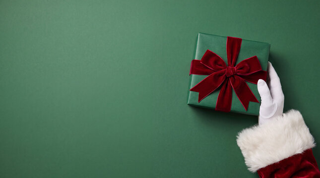 Santa Claus gloved hand placing a green Christmas gift box tied with a red velvet ribbon on a matching green background with copy space. High angle top view shot.