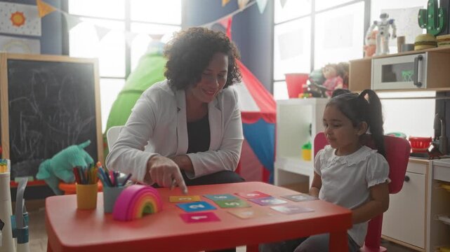 Woman teacher points finger to colorful flashcards on a red kids table in a playroom inside a building; learning joy.