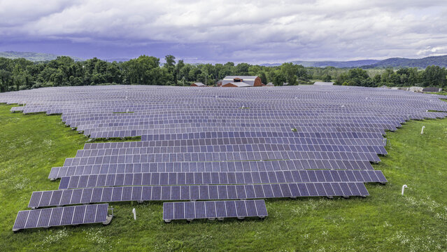 Aerial view of a large solar panel farm set in a green rural field with a red barn in the background under a cloudy sky in Patterson, New York, United States.