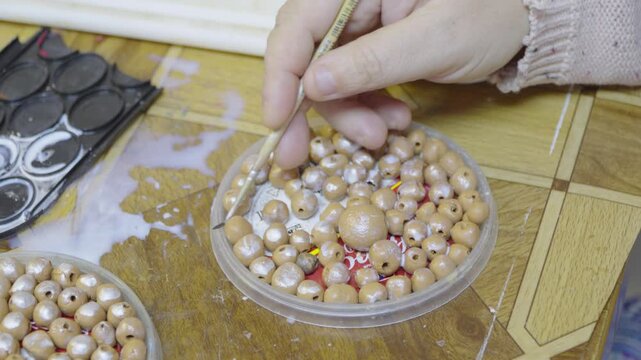 An artisan uses a paintbrush to carefully apply white paint onto small wooden beads arranged in a round container on a workshop table.