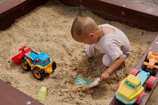 A young child is digging in a sandbox using a small shovel. Nearby, there are colorful toy trucks. The scene is in a playground during the daytime