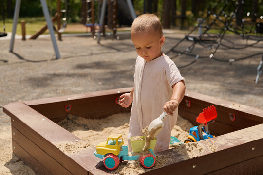 A young child plays in a sandbox, using a small truck to move sand. The playground is busy with swings and climbing structures in the background