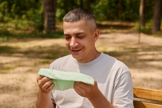A man sits on a wooden bench in a park, holding a cloth in his hands. He looks happy while gazing at the cloth. Trees surround the area, and it is sunny