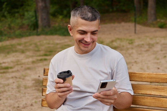 A man is seated on a wooden bench in a park. He holds a coffee cup in one hand and looks at his smartphone with a smile. The background is natural
