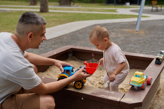 A father sits near a sandbox, helping his child with toys. The child uses a red shovel while playing in the sand. Nearby, colorful toy trucks are present