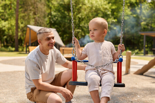 A man watches his small child on a swing in a park. The child sits quietly, and the man smiles as they enjoy the sunny day outdoors