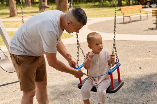 A man adjusts a swing for a young child at a playground. They are surrounded by trees and benches. The sun is shining and it's a warm day