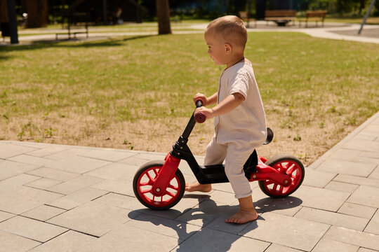 A young child balances on a small bike while pedaling across the pavement in a park. Green grass and trees surround the area on a warm day