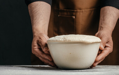 Fototapeta premium Male baker in brown apron holds a bowl of risen dough with a smooth surface, preparing for the next step in the baking process on a kitchen countertop