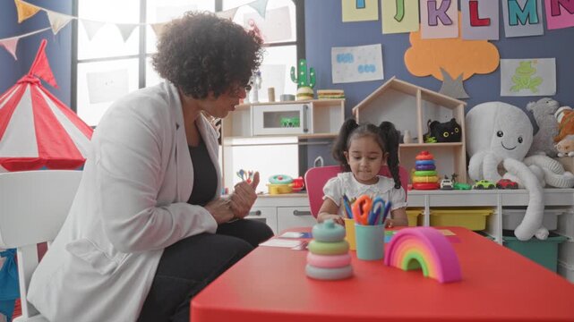Woman shows small toy with hand to little girl in preschool building while seated at a red table with stacking rings; playful learning.