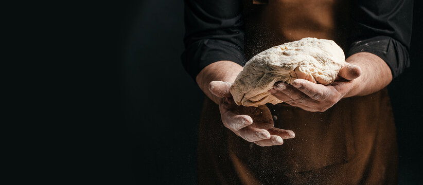 Male baker in brown apron holds kneaded dough with flour dusting in hands, dark background emphasizing the texture and details of the bread preparation process