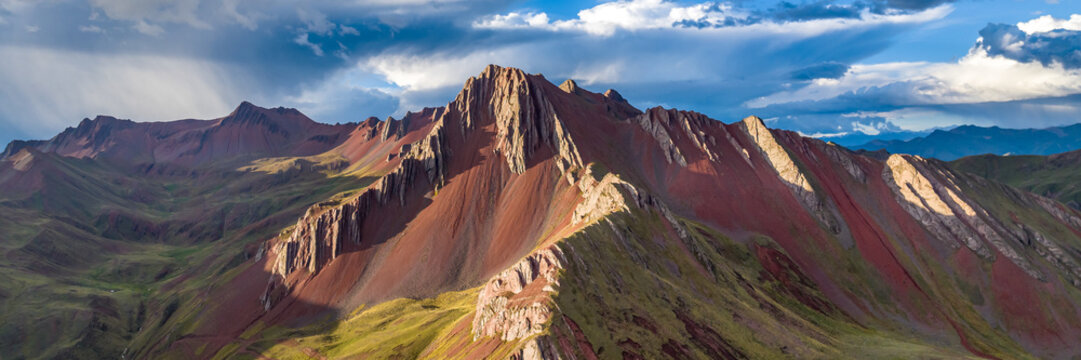 Aerial view of the colorful mineral-rich slopes of Vinicunca, featuring dramatic red and green mountain ridges under a cloudy sky in Quispicanchi Province, Cusco, Peru.
