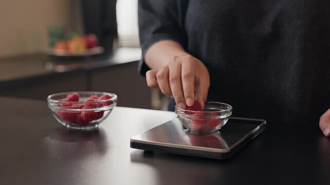 Young adult woman weighing fresh raspberries on digital kitchen scale, then eating one and tasting it thoughtfully. Perfect for healthy eating, nutrition apps, and diet tracking videos