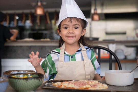 Smiling little girl wearing a chef hat and apron proudly showing homemade pizza during a children's cooking class. Hands-on culinary education encourages creativity, confidence, learning, family fun.