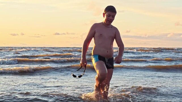A boy comes ashore from the ocean after swimming with swimming goggles in his hand. Sports and recreation on the water