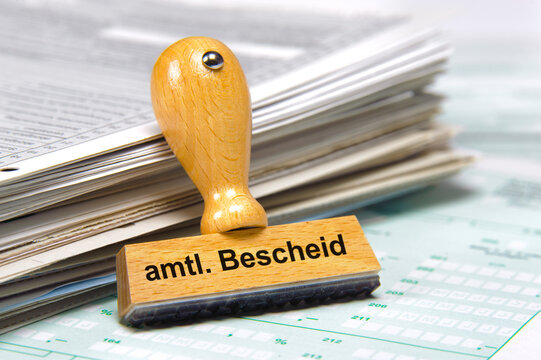In einem B&uuml;ro lehnt ein Holzstempel an einem Stapel Papiere mit der Aufschrift: amtl. Bescheid