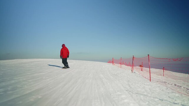 A lone snowboarder in a vibrant red jacket descends a pristine snow-covered mountain slope. The adventurous individual enjoys a thrilling winter sport against a clear blue sky.