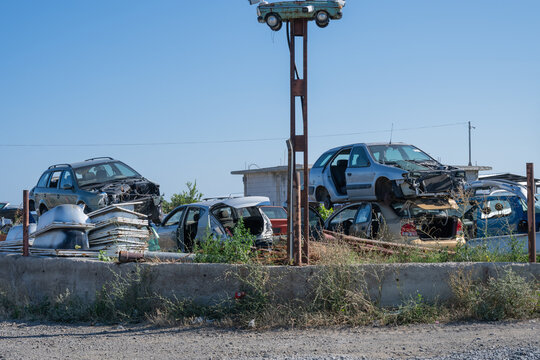 an auto salvage yard featuring stacks of dismantled vehicles behind a low concrete wall, with a small car mounted high on a rusty pole acting as a display sign.