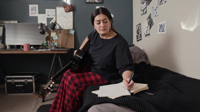 Young adult woman playing acoustic guitar in bedroom, then writing lyrics in notebook. Ideal for music education, creative hobby, or artistic lifestyle commercials