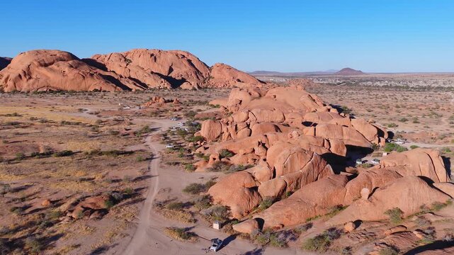 Aerial view of Spitzkoppe inselbergs in Erongo, Namibia. Rounded orange brown domes rise from desert. Natural arches, tracks, and campsites show visitor activity.
