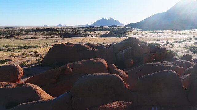 Aerial drone pans and descends over Spitzkoppe in Erongo, Namibia. Dirt tracks, shrubs, and tiny vehicles appear. Low sun casts long shadows and lens flare.