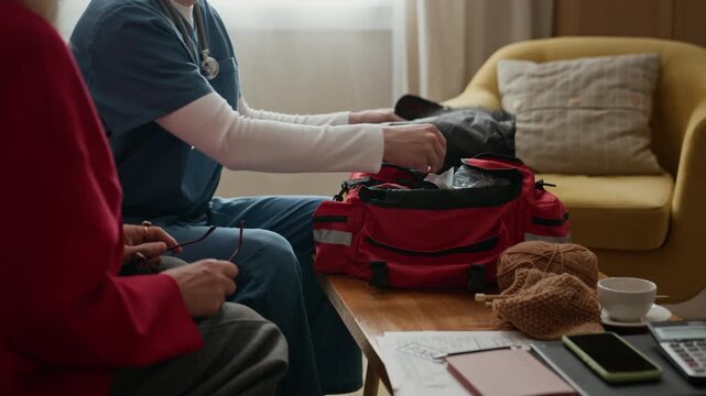 Young Caucasian male nurse opening red medical bag to retrieve blood pressure monitor while sitting next to senior Caucasian woman. Footage is ideal for home caregiving and geriatric medical services