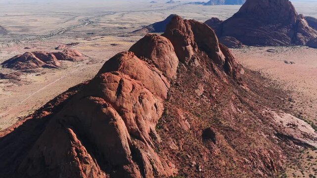 Aerial view of Spitzkoppe in central Namibia shows rust red domes, a serrated ridge, talus slopes, desert scrub, pale tracks, and a braided dry riverbed in bright light.