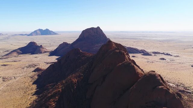 Aerial drone shot sweeps along Spitzkoppe inselbergs in Namibia, main dome and koppies in crisp daylight, wide high angle, barren plains to a clear horizon.
