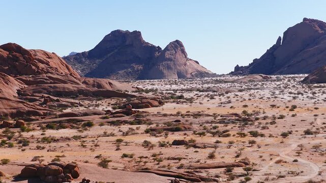 Aerial daylight view of Spitzkoppe in Namibia shows red brown inselbergs, smooth domes, jagged peaks, shrubs, tracks, and a lone white 4x4 moving across the sandy plain.
