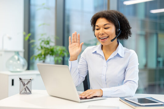 Smiling young African American businesswoman in office and headset holding online meeting, greeting laptop camera