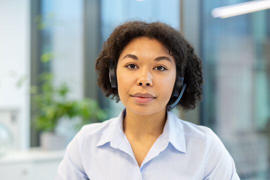 Portrait of a young African American woman in a headset sitting at a desk in the office and looking seriously at the camera