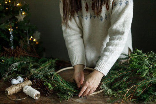Woman in cozy sweater making Christmas wreath on wooden table near decorated xmas tree. DIY holiday workshop in festive scandinavian interior with garland lights on tree