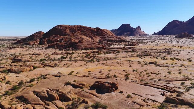 Aerial view of Spitzkoppe inselbergs and sandy plains in Namibia. A drone glides past rounded granite, salt flats, shrubs, and a tiny campsite at midday.
