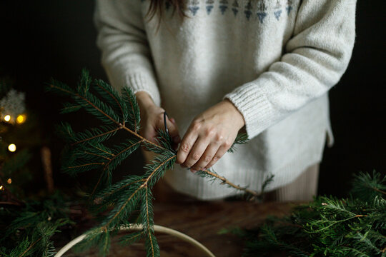 Female hands cutting fir branch with scissors to make Christmas wreath on rustic wooden table,close up. Florist workshop process, winter holiday decoration