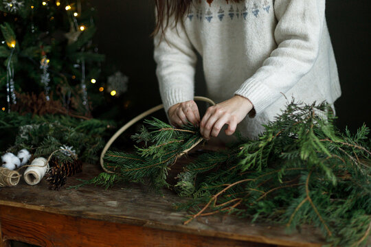 Making christmas rustic wreath. Female hands holding fir branches for handmade Christmas wreath in front of glowing xmas tree. Cozy winter holiday preparation