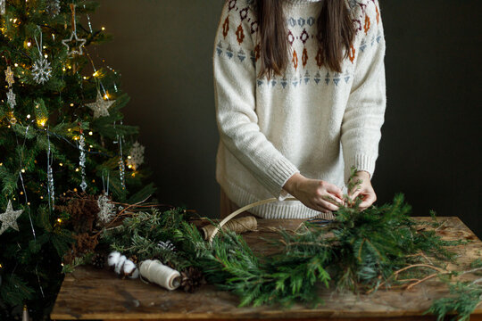 Woman in cozy sweater making Christmas wreath on wooden table near decorated xmas tree. DIY holiday workshop in festive scandinavian interior with garland lights on tree