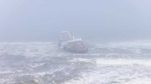 Aerial view shows a grounded trawler on Namibia Skeleton Coast, listing in heavy surf. Waves break, foam and spray swirl, fog mutes tones as the camera drifts overhead.