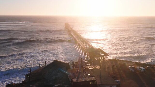 Aerial view shows a long stilted jetty on Namibia's Atlantic coast at sunset. Waves roll and break, cars park on the headland, flags fly, people gather, and lens flare reflects.