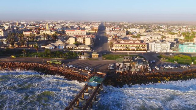 Aerial view of Swakopmund, Namibia, shows the pier, rocky breakwater, palm promenade, German colonial buildings, cafes, and moving people and cars in warm golden hour light.