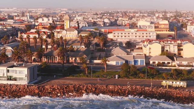 Aerial view of Swakopmund, Namibia shows palm promenade, rocky seawall, red breakwater, clock tower, town center, parked cars, bus at stop, waves and people strolling.