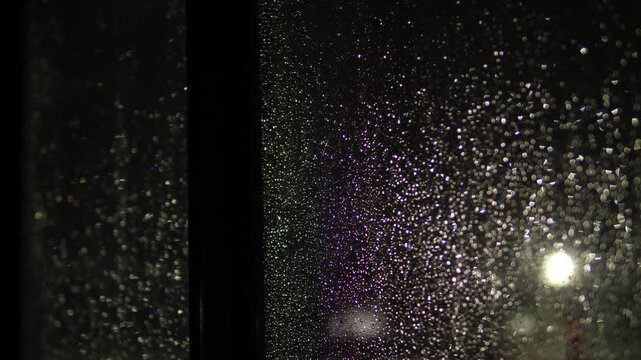 Small raindrops cover the window frame glass and shimmer with purple-green glints under night lighting. The blurred divider and fuzzy street lights create deep bokeh and a cozy cinematic atmosphere